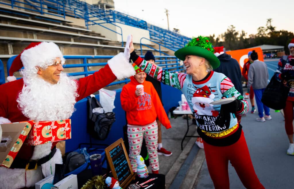 Santa Claus in a red suit high-fives a woman in a festive Christmas sweater and tree-shaped hat at a Gainesville Run Club event near stadium seating.