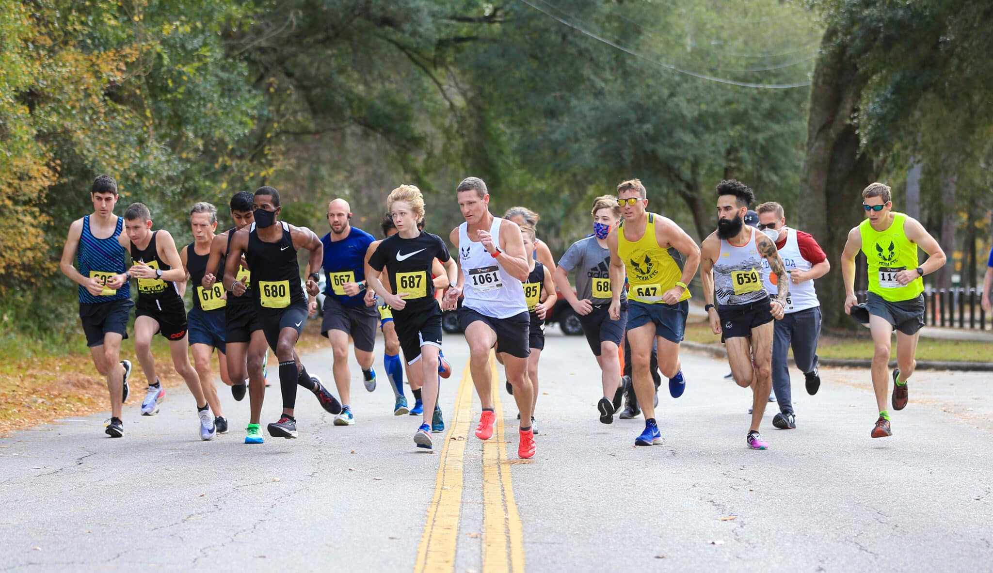 A group of Florida Track Club runners wearing race bibs sprint down a paved road during a race.