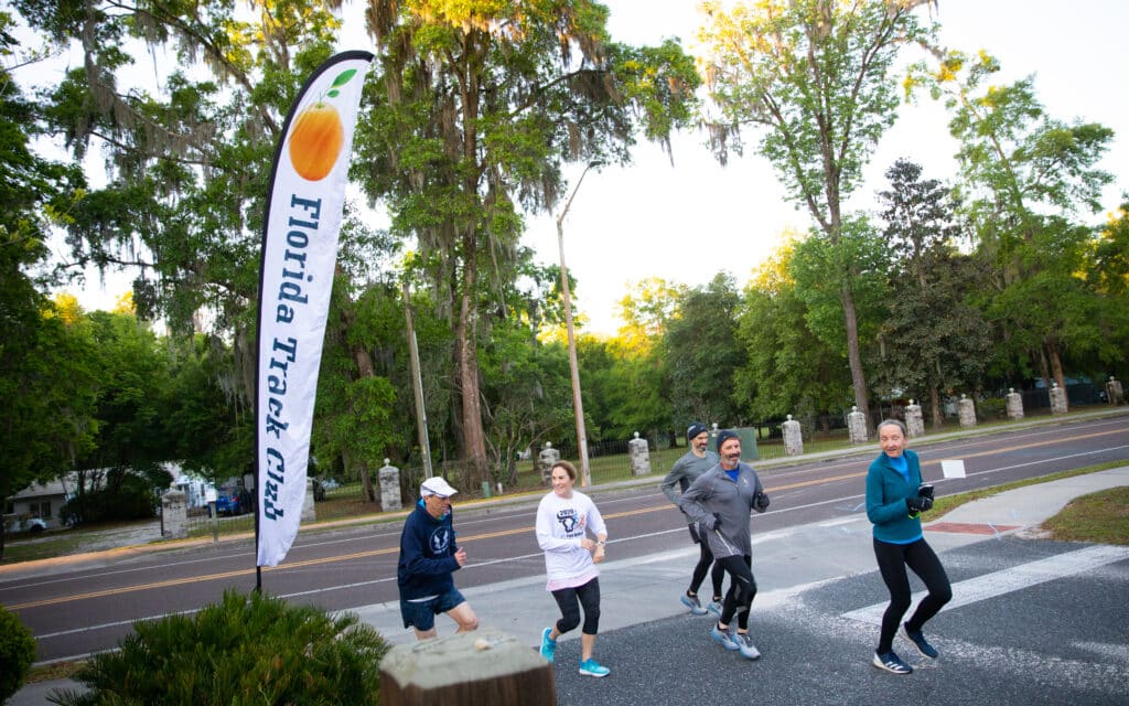 A group of runners from the Florida Track Club jogs past their banner in a park.