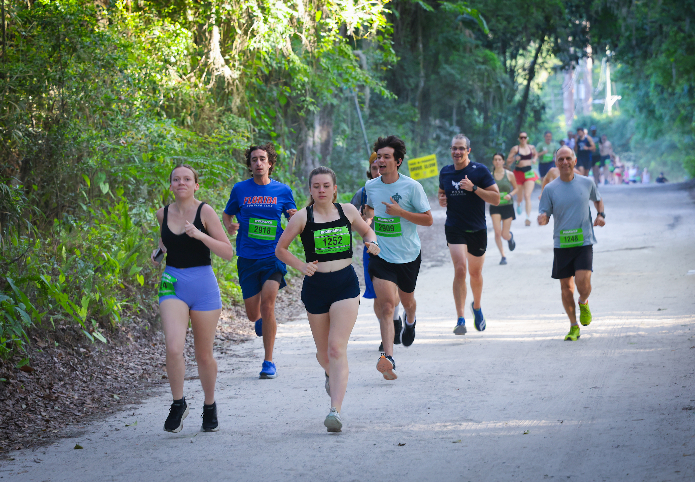 Members of the Gainesville Run Club wearing race bibs jog along a dirt trail lined with trees.