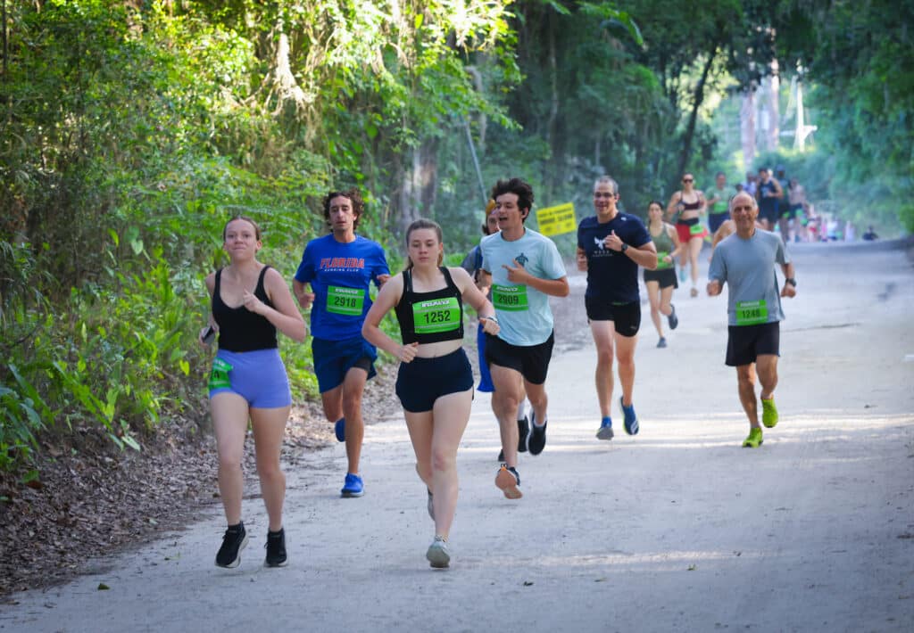 A group of Gainesville Running Club members in athletic wear participating in a race along a dirt path through a wooded area.