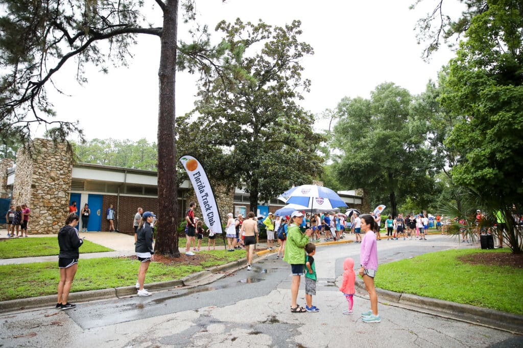People gather in a park during a rainy day for a Florida Track Club event, with several participants holding umbrellas.