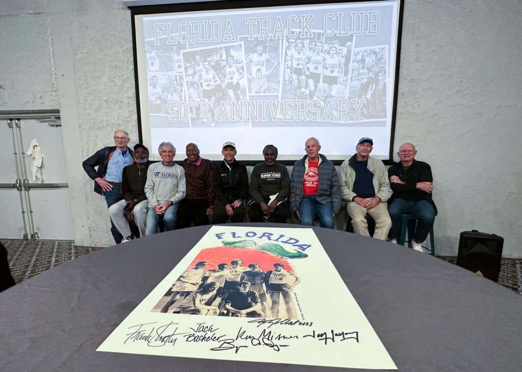 Eight men sit in a row before a projection screen displaying a Florida Track Club 50th anniversary poster, with a matching signed poster resting on the table in the foreground.