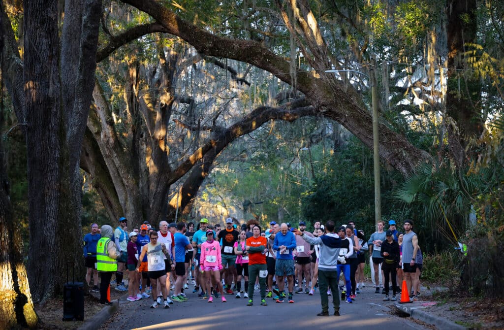 A group of runners wearing race bibs from the Gainesville run club stand on a paved road under large, moss-draped oak trees while a man in a vest gestures toward them.