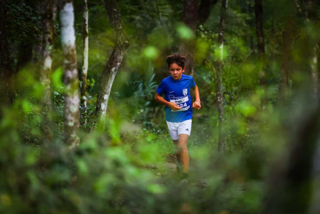 A young boy wearing a blue shirt with the race number 224 runs along a wooded trail during a Gainesville running club event.