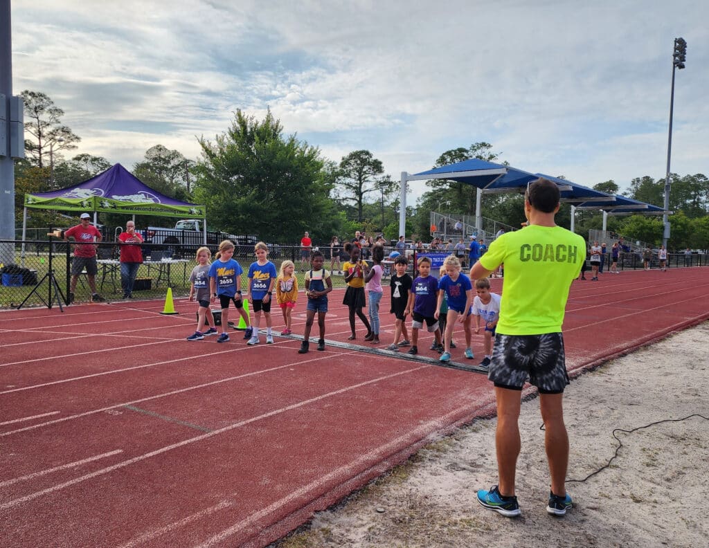 A Florida Track Club coach in a neon yellow shirt stands on a track facing a line of young children prepared to start a race.