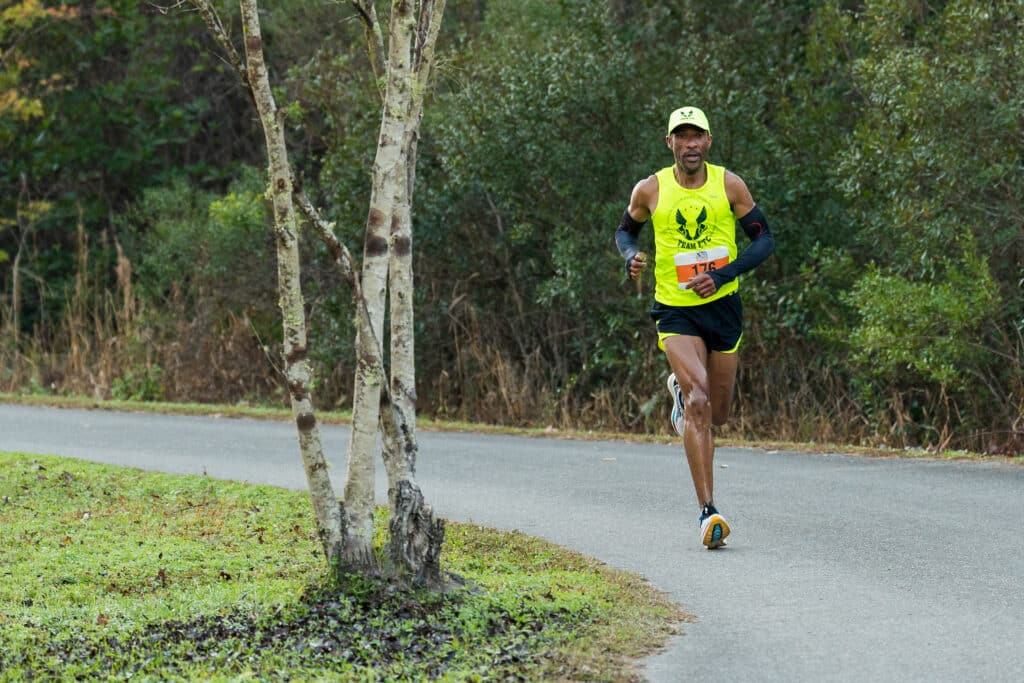 A male runner from the Gainesville Running Club wearing a neon yellow tank top, black shorts, and a race bib runs along a paved path in a wooded area.