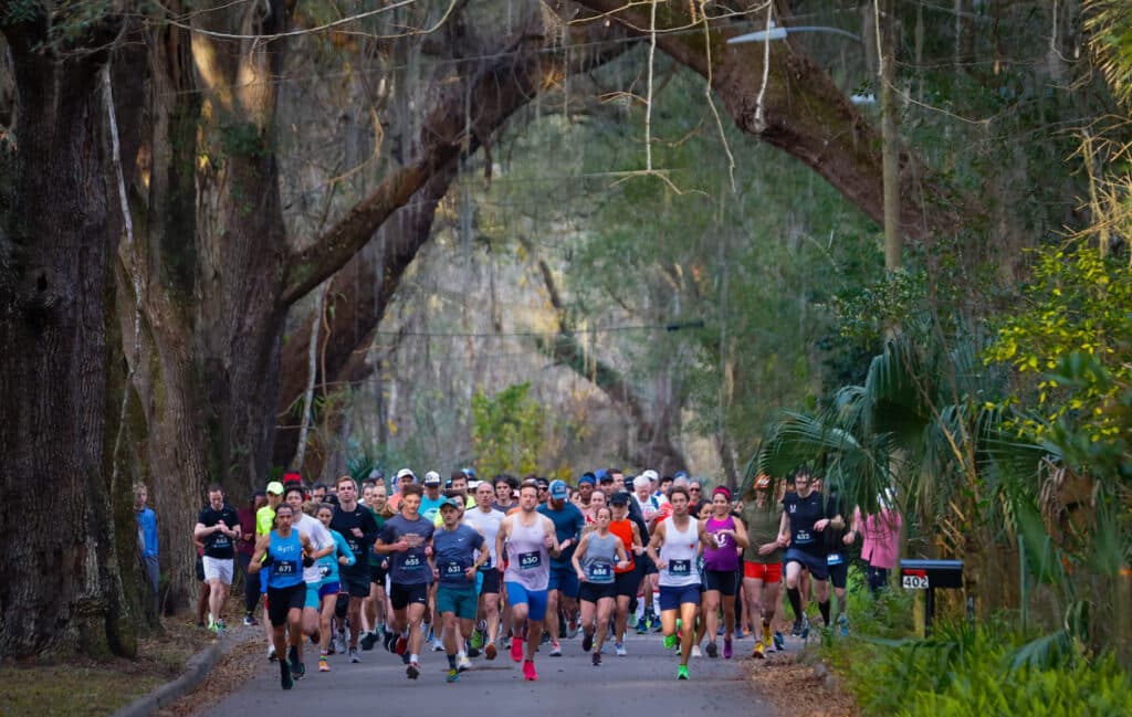 A large group of Gainesville Running Club members participating in a road race on a paved path lined with large oak trees.