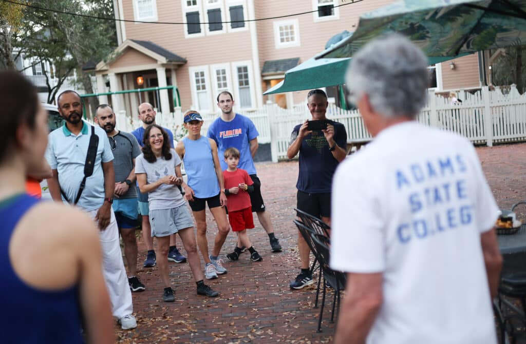 A group from the Florida Track Club stands on a brick patio listening to a man in an Adams State College t-shirt, while another man photographs them with a phone.