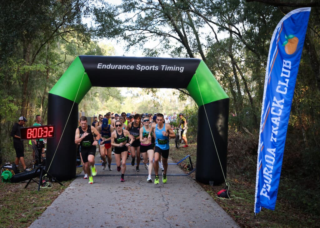 Runners cross the finish line under an “Endurance Sports Timing” arch with a race clock showing 8:00:32 and a Gainesville Running Club banner nearby.