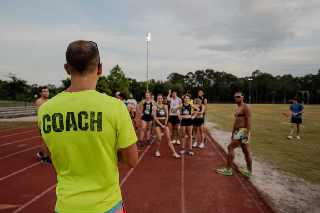 A coach in a neon yellow shirt with “COACH” printed on the back faces a group of runners from the Gainesville Running Club on a track.