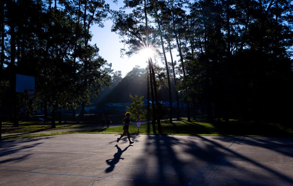A silhouette of a Florida Track Club member running across an outdoor basketball court with the sun shining through tall trees in the background.