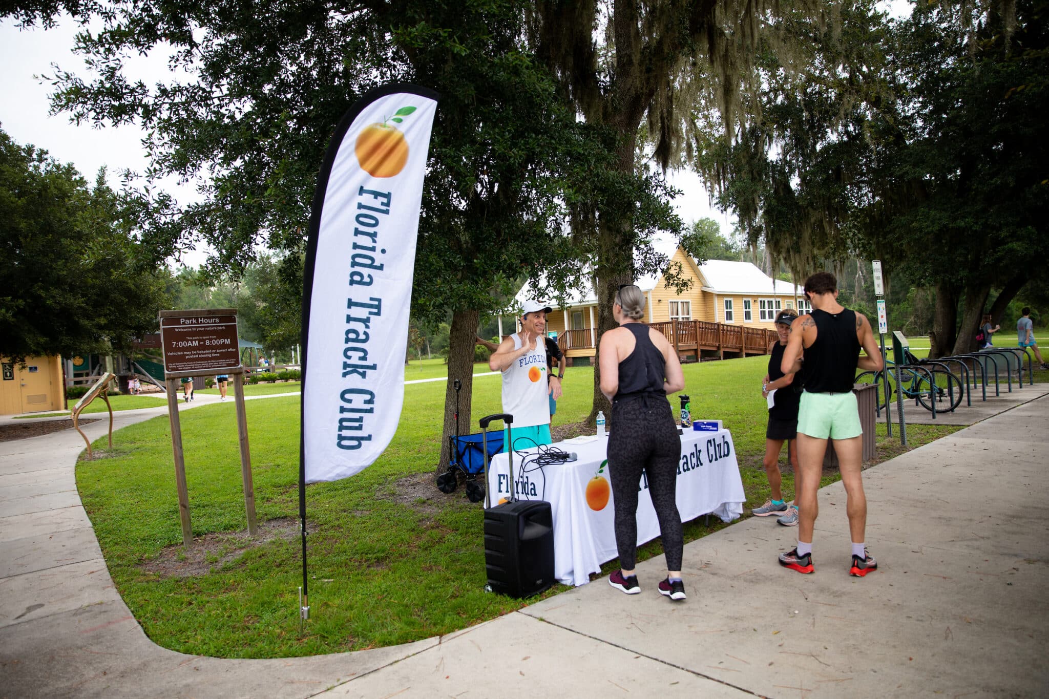 Three people stand at a Florida Track Club information table on a sidewalk near a park.