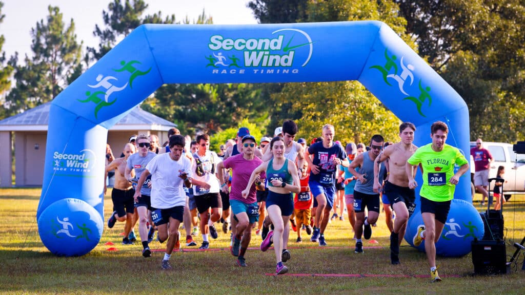 A group of Florida Track Club runners in athletic wear passes under a large blue inflatable arch labeled “Second Wind Race Timing” at the start of a race.