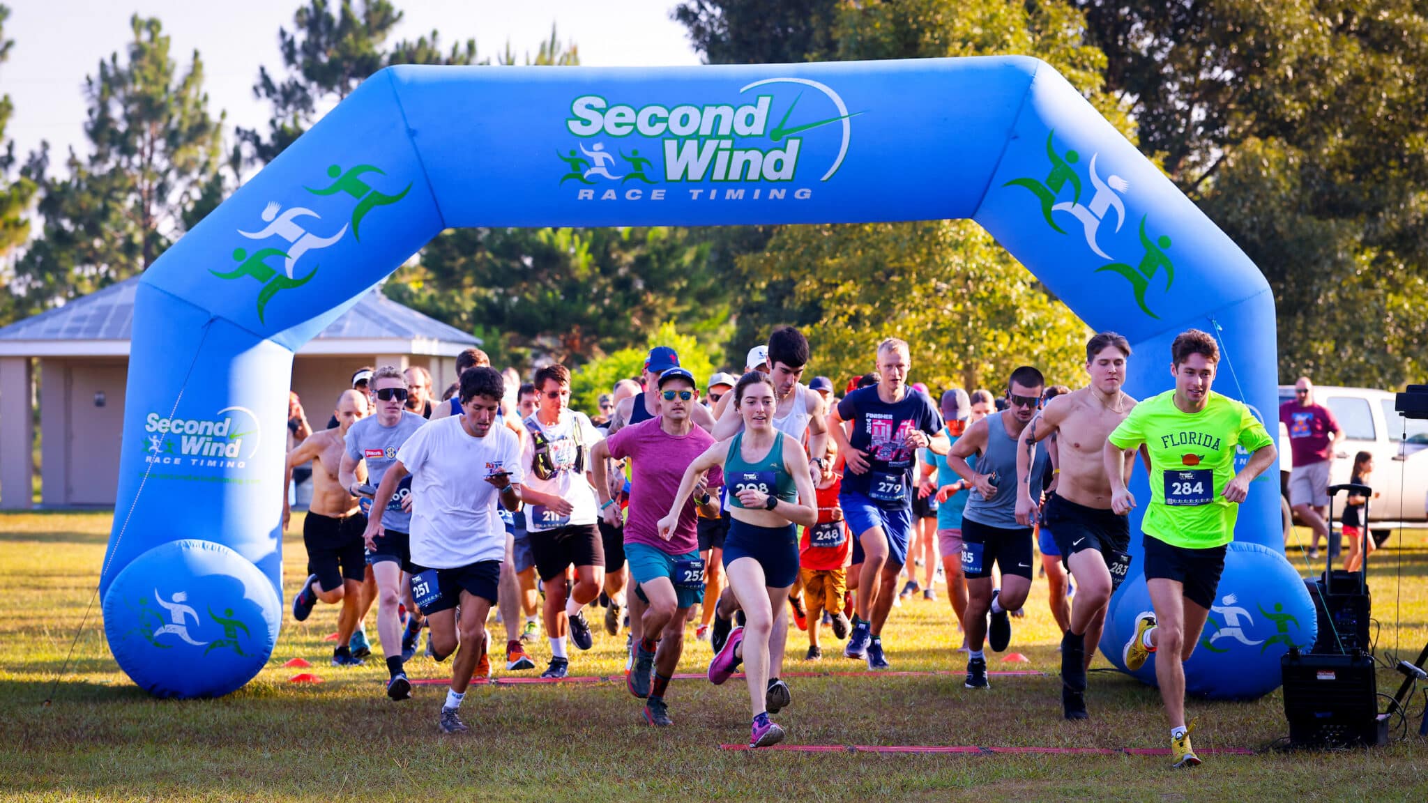 A group of Florida Track Club runners in athletic wear passes under a large blue inflatable arch labeled “Second Wind Race Timing” at the start of a race.