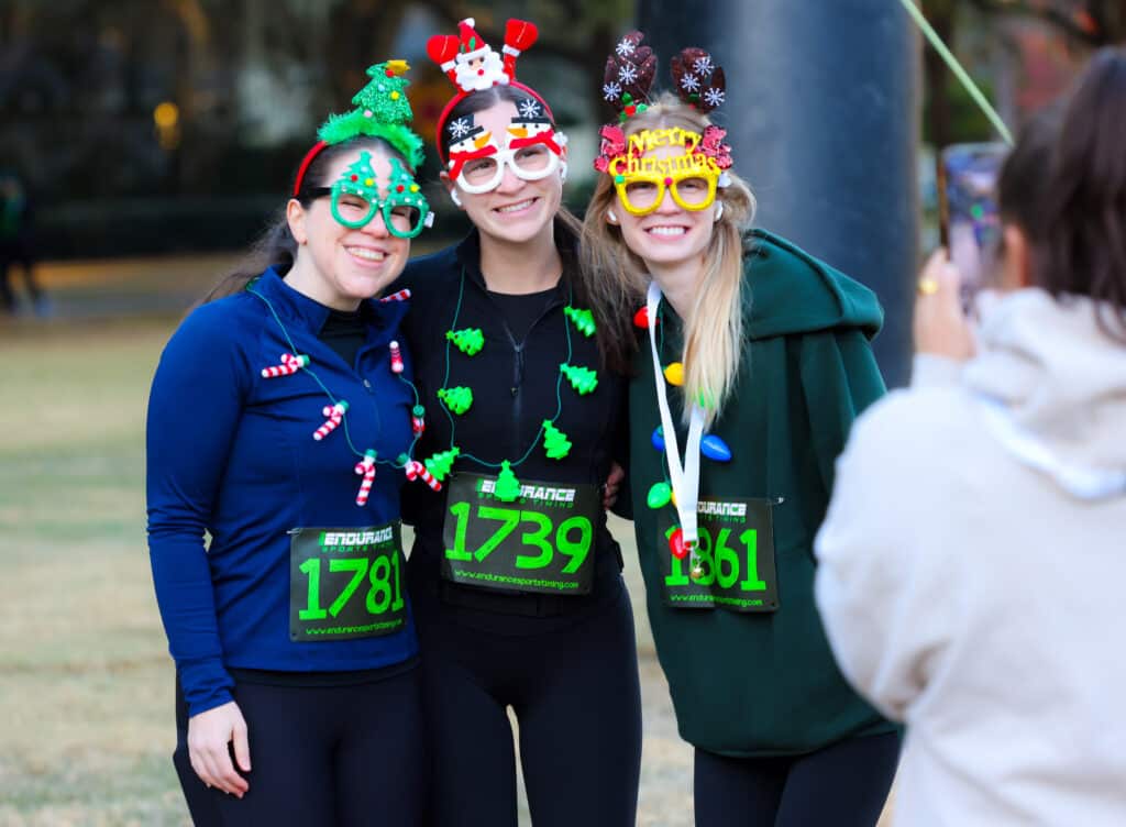 Three Florida Track Club members wearing festive Christmas headbands and glasses pose for a photo while wearing race bibs.