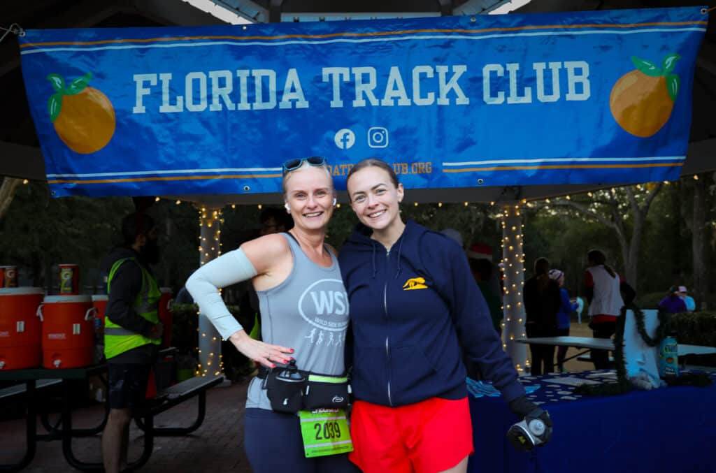Two women posing in front of a Florida Track Club banner at an event.