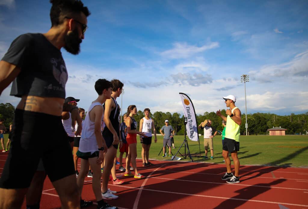 A coach in a yellow tank top speaks to a group of athletes on a running track near a Florida Track Club banner.
