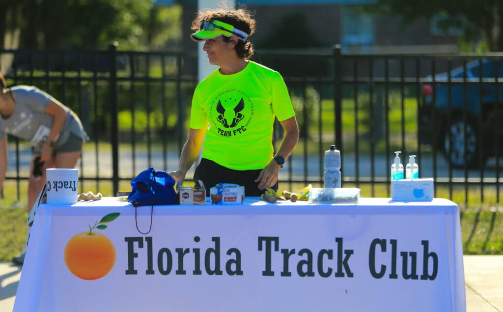 A runner in a neon green shirt stands behind a table with a Florida Track Club banner, with various supplies laid out for this Gainesville running club.