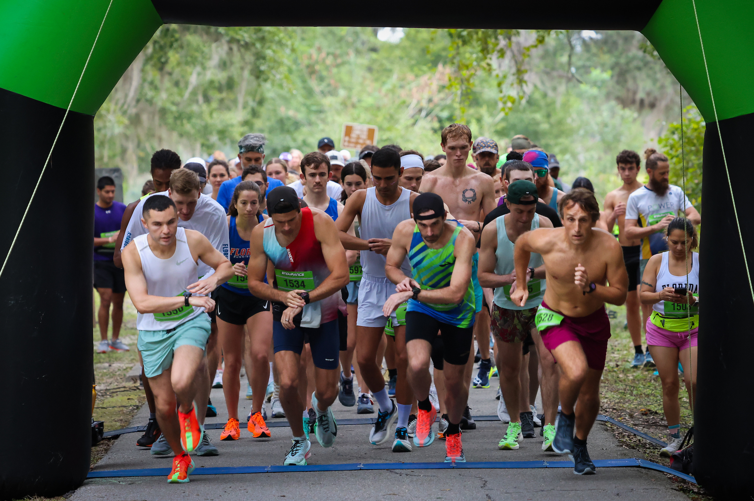 A large group from the Gainesville Run Club starts a race, passing through a green inflatable archway while checking their watches.