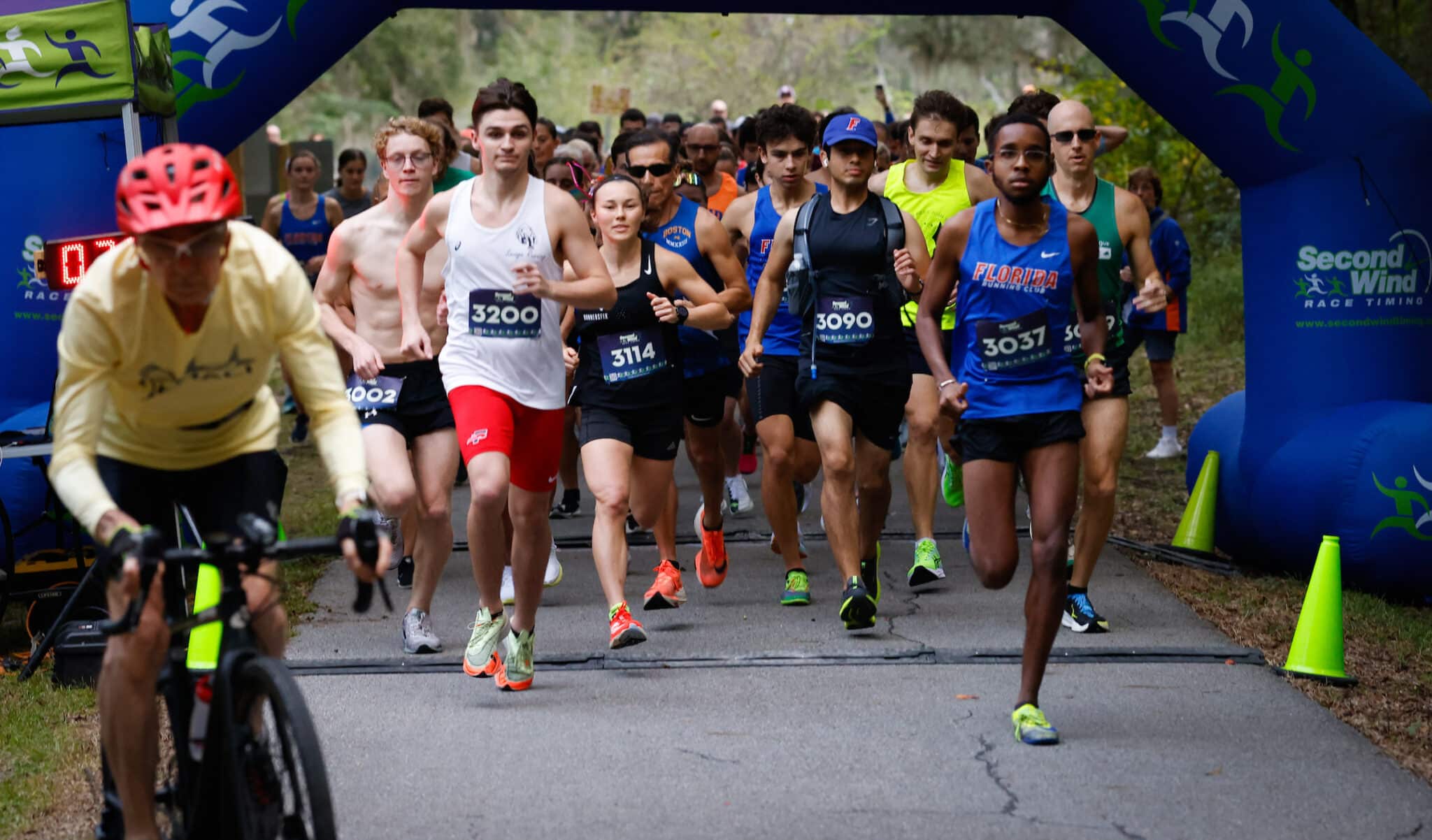 A group of Gainesville Run Club members wearing race bibs sprint toward the finish line under a blue inflatable archway.