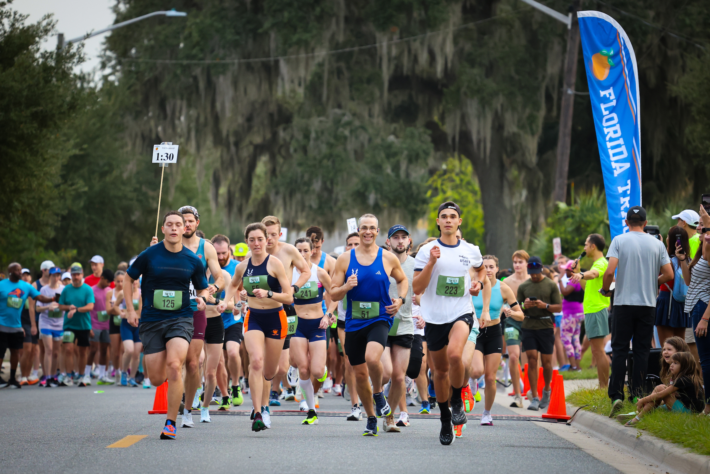 A group of Gainesville Running Club members participates in a race on a paved road, led by a pack of front-runners wearing bibs and athletic gear.