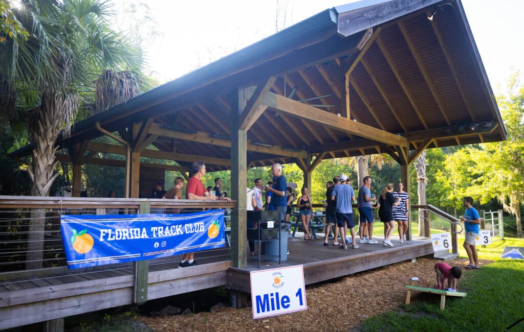 Florida Track Club members socialize on the deck of a wooden pavilion, with mile marker signs displayed nearby.