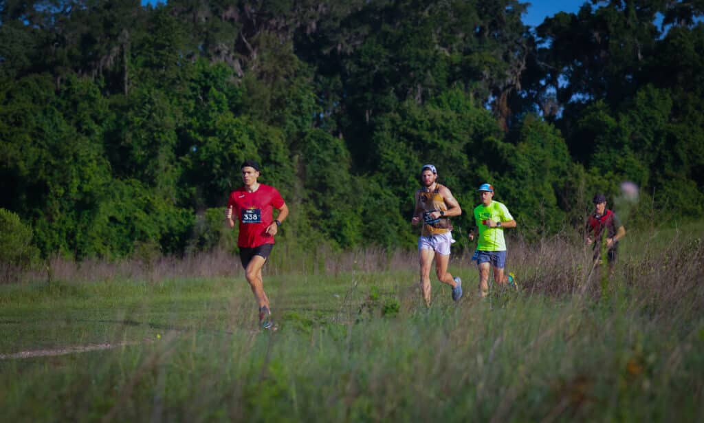 Four runners from the Gainesville Running Club racing along a grassy trail with trees in the background.