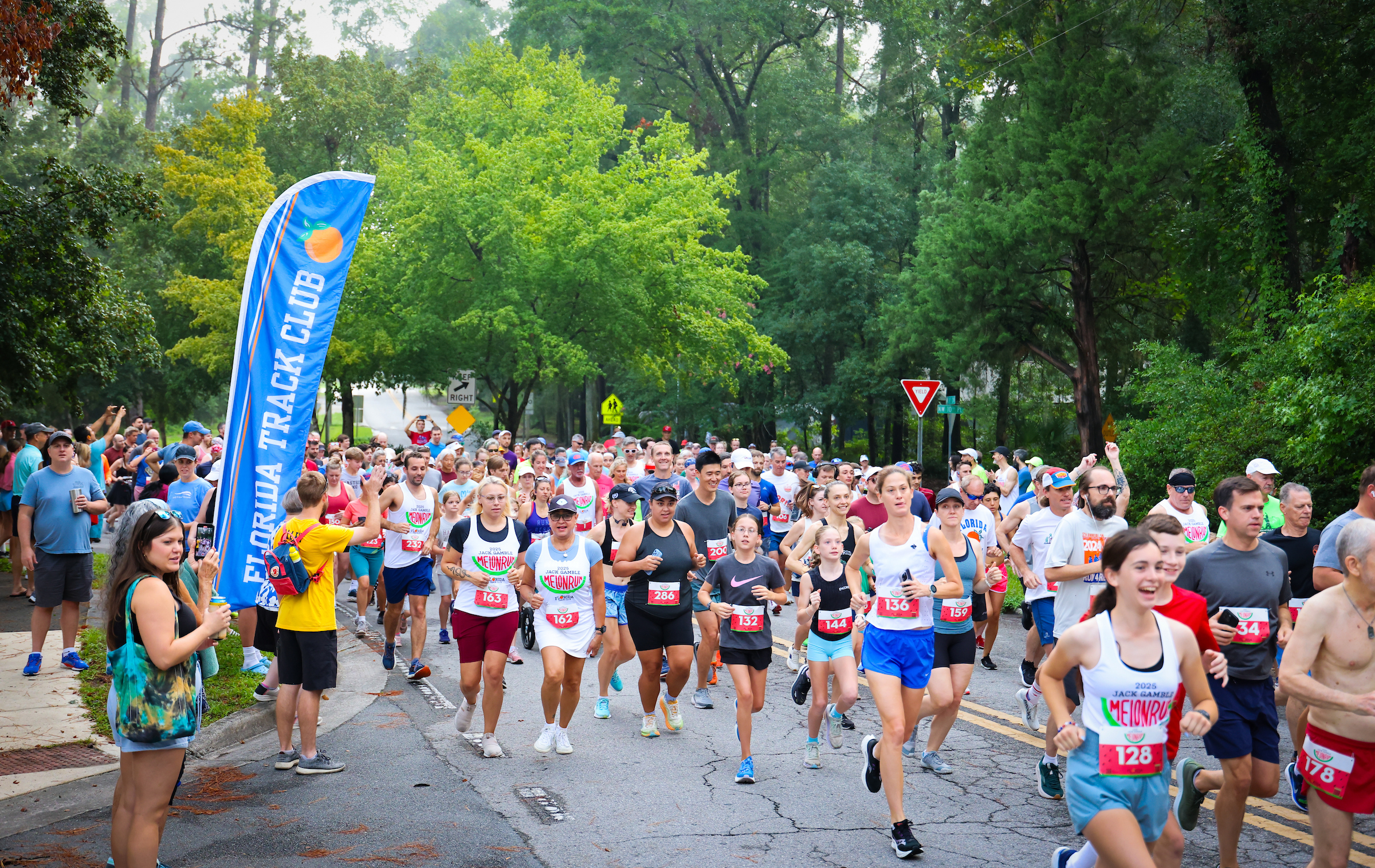 A large group of runners participates in the 2024 Jack Gamble Melon Run on a tree-lined street, passing a Florida Track Club banner on the left.