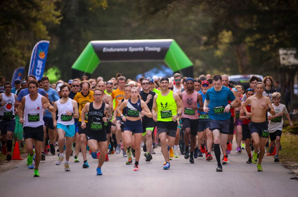 A large group from the Gainesville Run Club participates in a road race, heading toward a green “Endurance Sports Timing” archway.