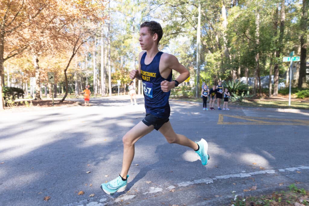 A male runner from the Gainesville Run Club wearing a navy tank top with “Distance Project” and bib number 67 runs along a paved road.