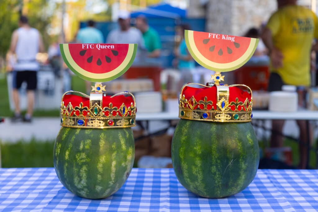Two watermelons wearing decorative crowns and signs labeled “FTC Queen” and “FTC King” sitting on a blue checkered tablecloth at a Gainesville running club event.