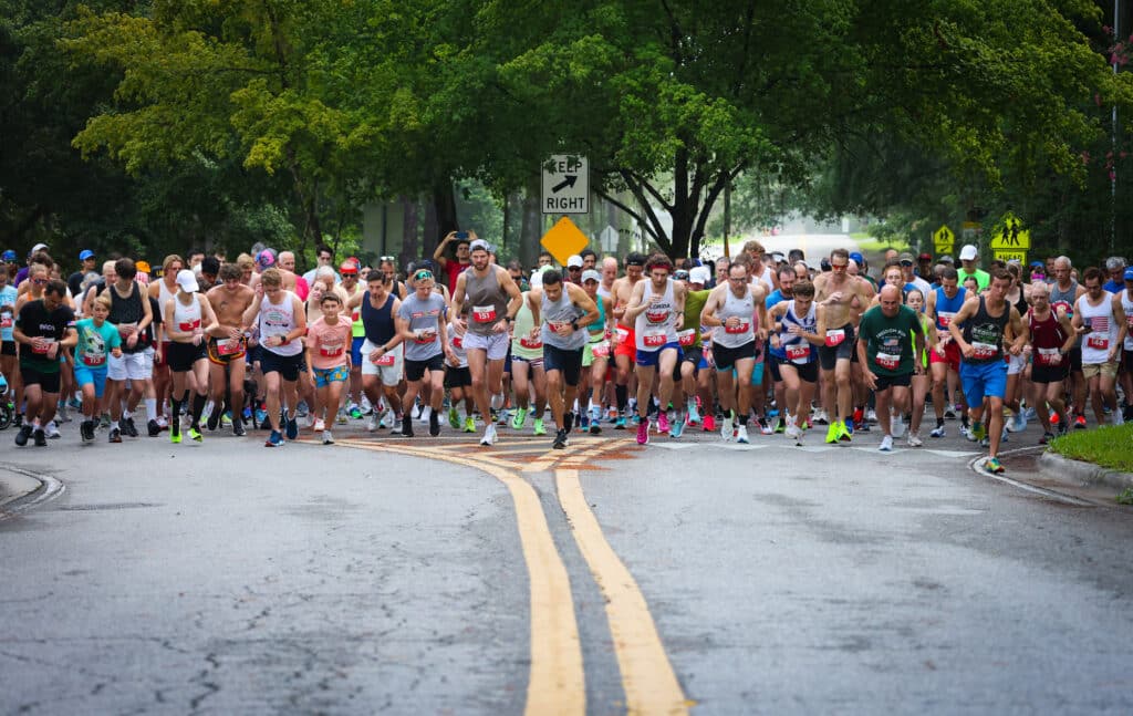 A large group of runners from the Gainesville Running Club wearing race bibs begins a road race, heading toward the camera on an asphalt street.