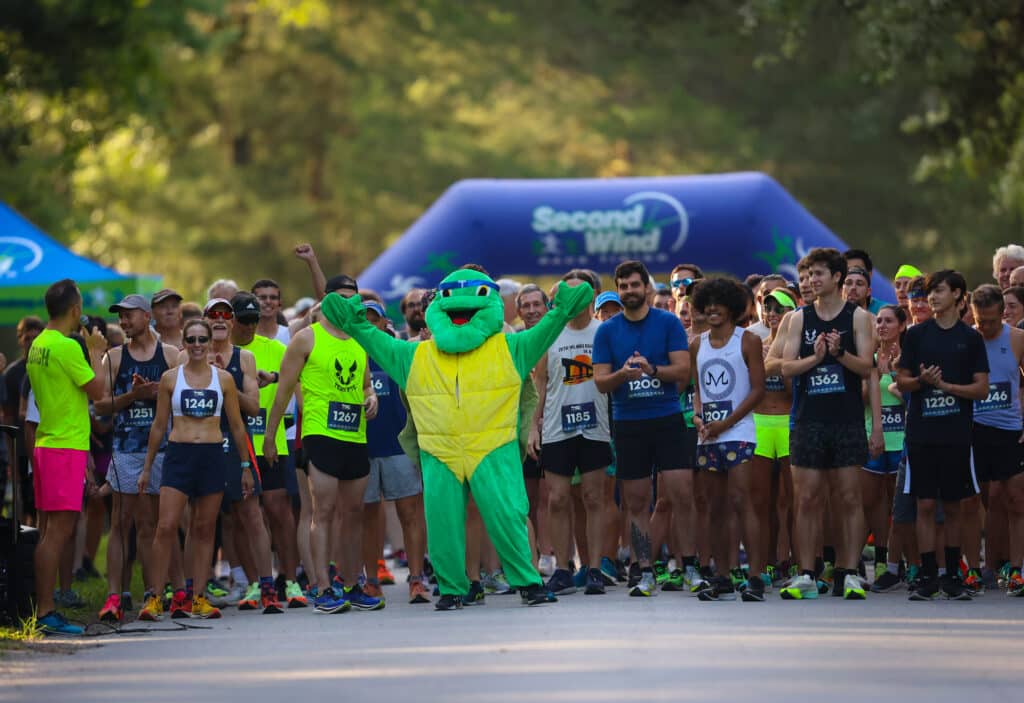 A person in a turtle mascot costume stands with arms outstretched in front of a crowd from the Gainesville Run Club at a race starting line.