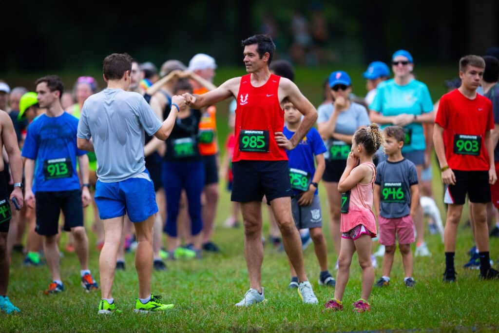 Two members of the Gainesville Running Club fist-bump at a race start line as other participants wait nearby.