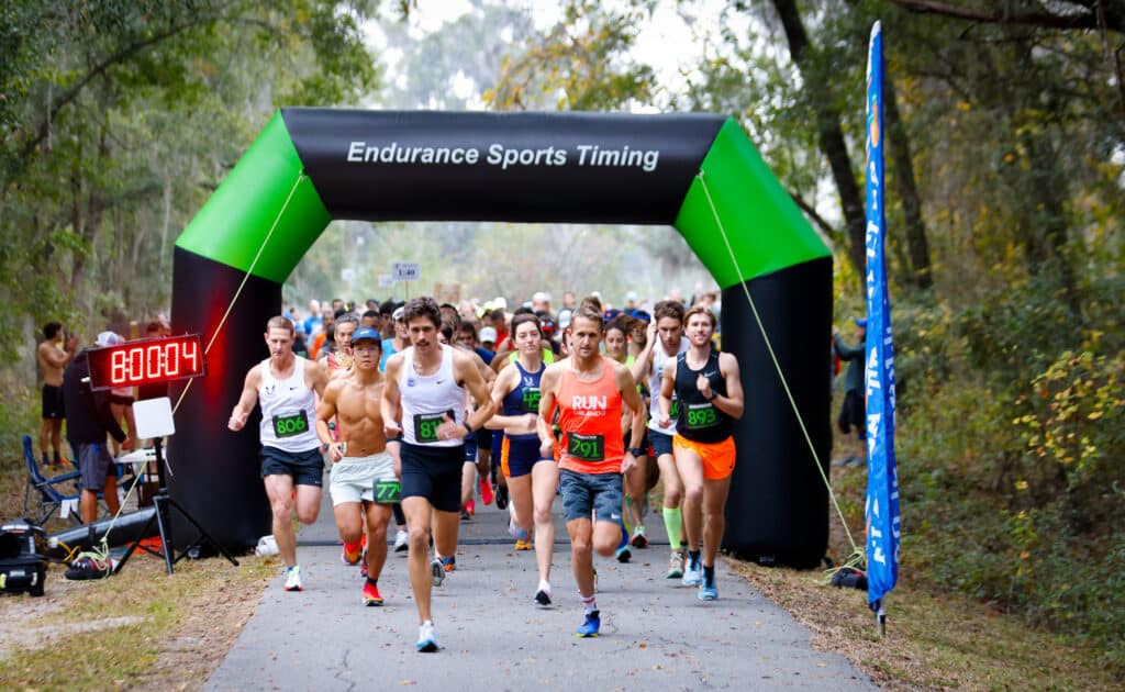 Runners from the Florida Track Club start a race beneath an “Endurance Sports Timing” inflatable arch with a digital clock displaying 8:00:04.