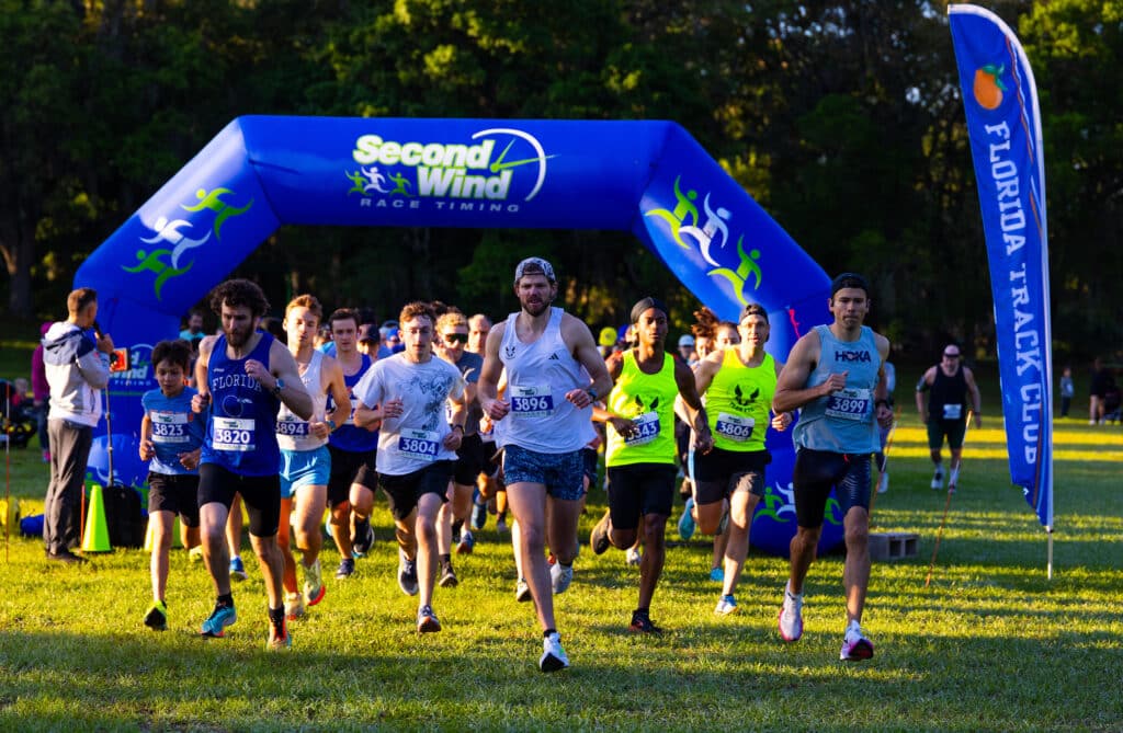 A group of runners from the Florida Track Club wearing race bibs sprint toward the finish line under a large blue inflatable arch labeled “Second Wind Race Timing.”