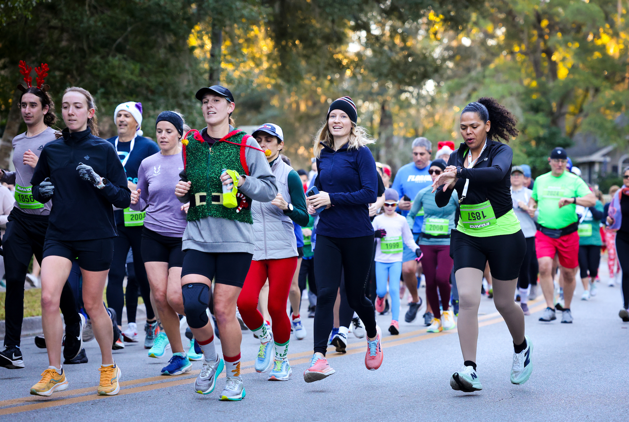 A diverse group from the Florida Track Club, including a woman in a festive elf costume, participates in a road race while wearing bib numbers.