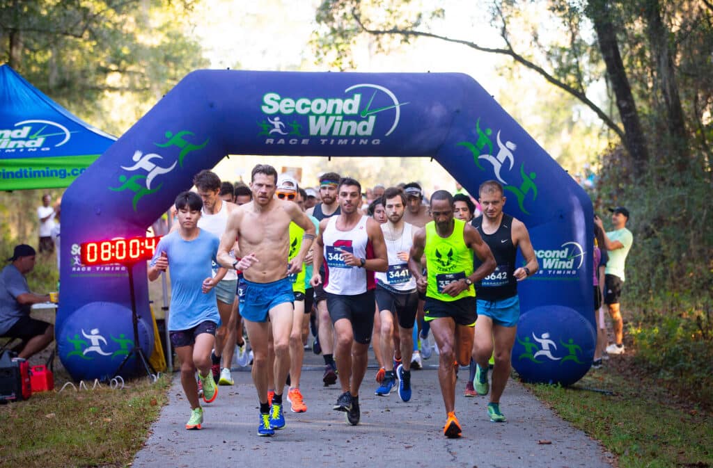 Runners in athletic wear compete in a race, passing through a blue inflatable finish line arch for the Florida Track Club displaying the Second Wind Race Timing logo and a digital race clock.