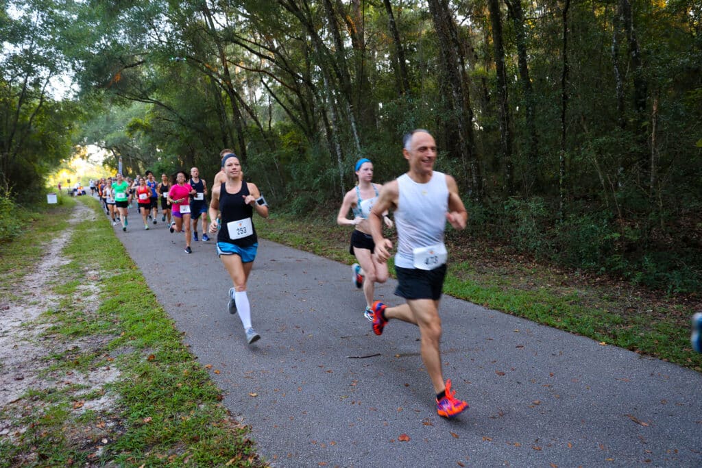Members of the Florida Track Club in athletic gear participate in a road race on a paved path lined with trees.