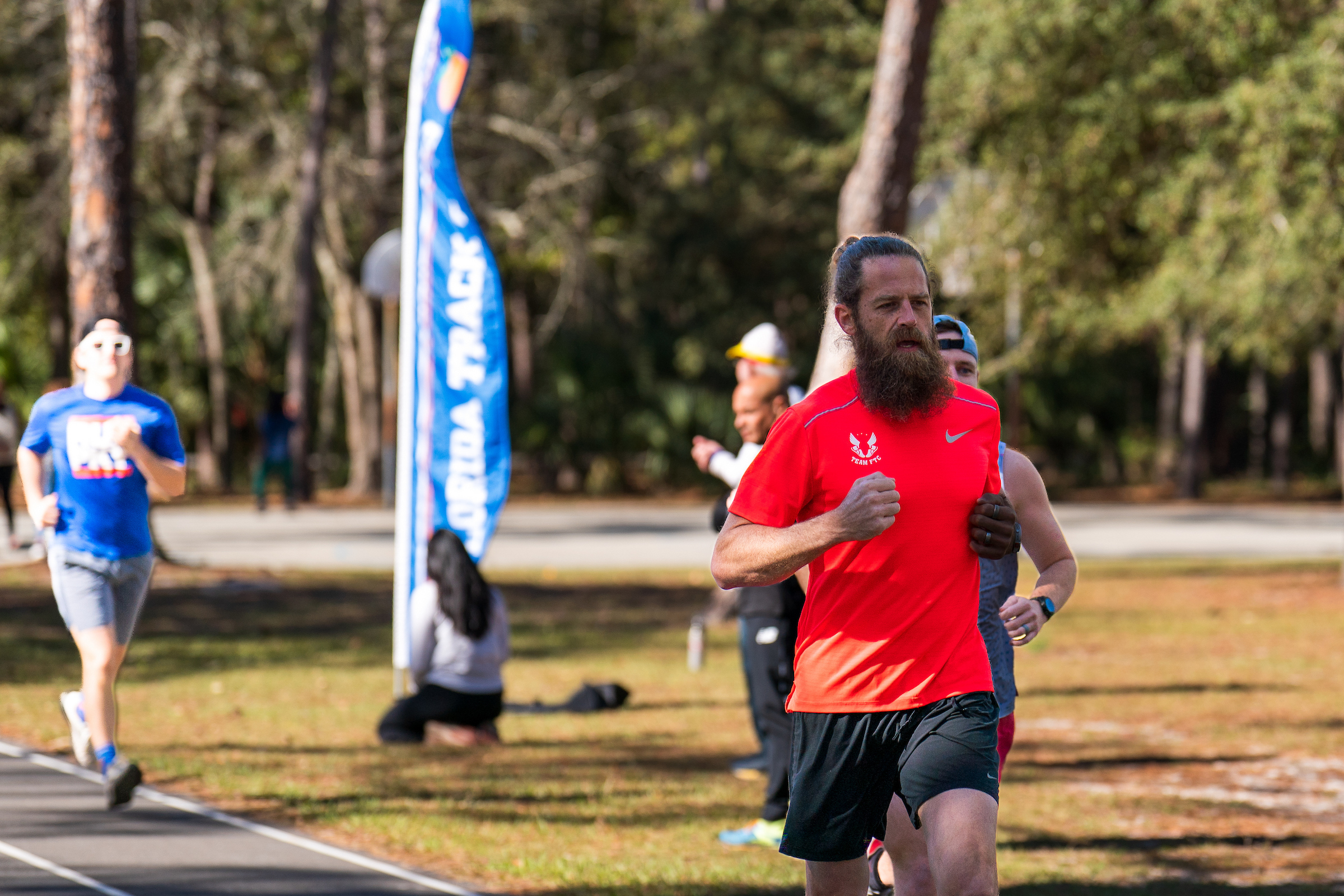 A bearded man in a red Gainesville Running Club performance shirt runs on a track during a race with other runners in the background.