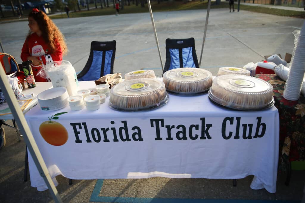 A Florida Track Club table at an outdoor event featuring stacks of plastic containers, paper plates, and several round catering platters from the Gainesville running club covered with clear plastic lids.