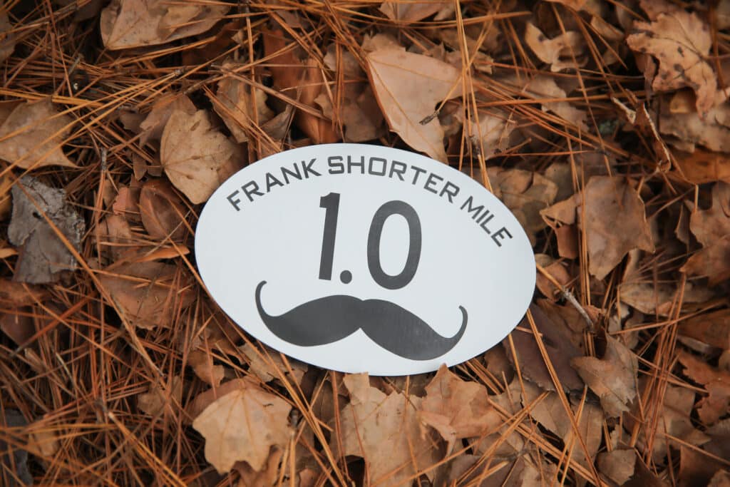 An oval race marker on a bed of dry leaves reading “Frank Shorter Mile 1.0” with a black mustache icon below the number, marking a route for the Florida Track Club.