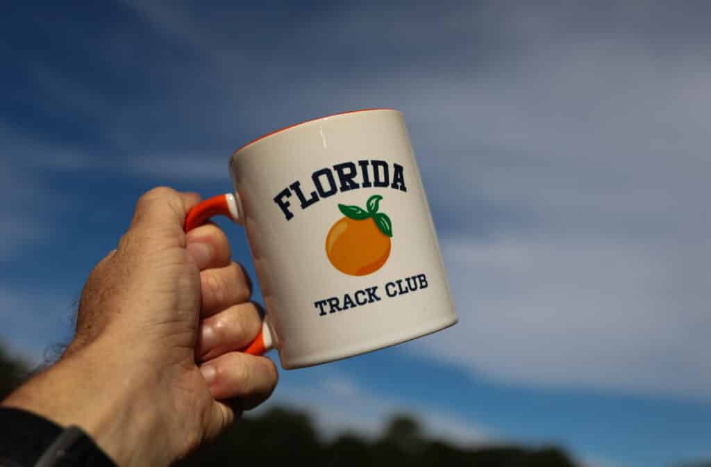 A hand holding a white mug with an orange Florida Track Club logo, a popular Gainesville running club.