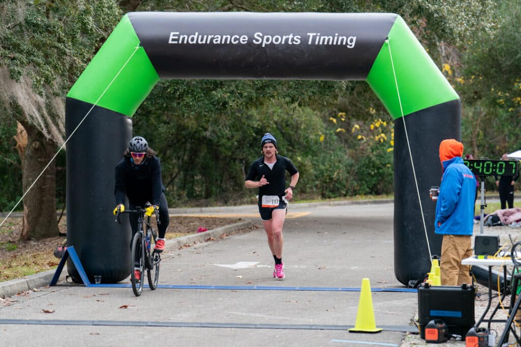 A cyclist and a runner pass under an “Endurance Sports Timing” archway at a Florida Track Club event.