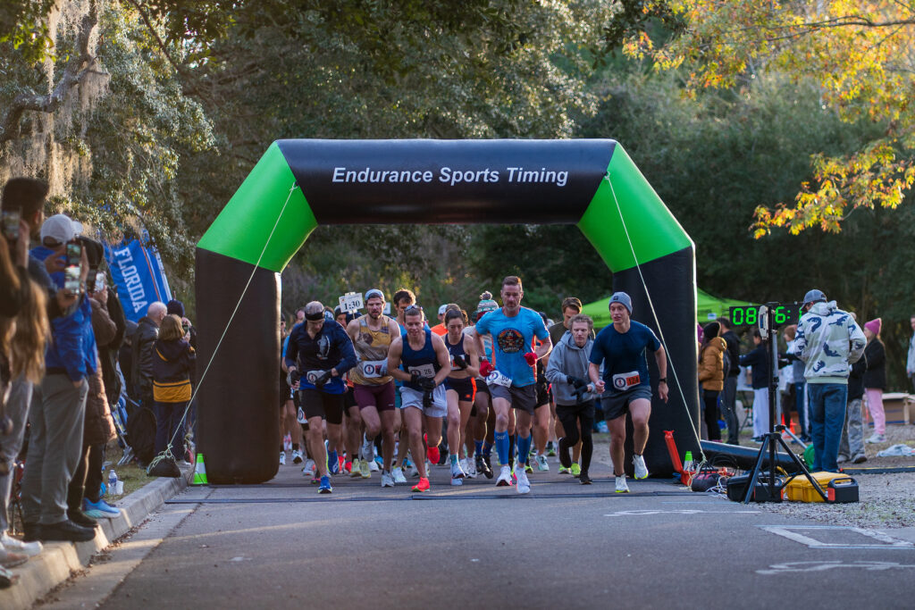 Runners from the Florida Track Club cross the start line of a race under an inflatable archway as spectators watch from the sidelines.