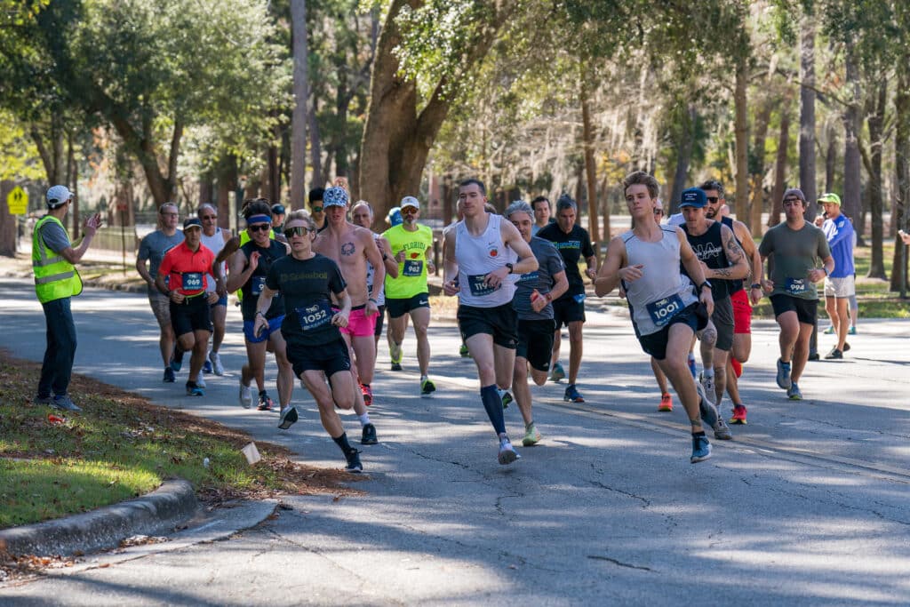 A group of Florida Track Club runners participates in a road race on a paved path lined with trees.