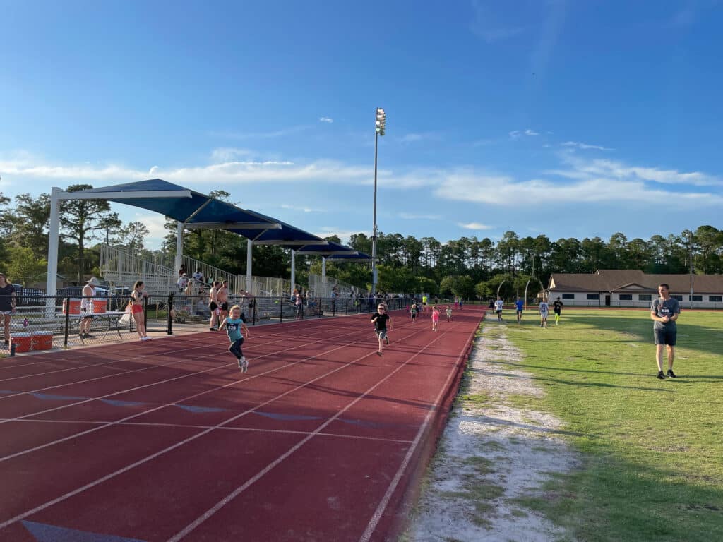 Children and adults participate in a Gainesville Run Club event on a red track at an outdoor stadium.