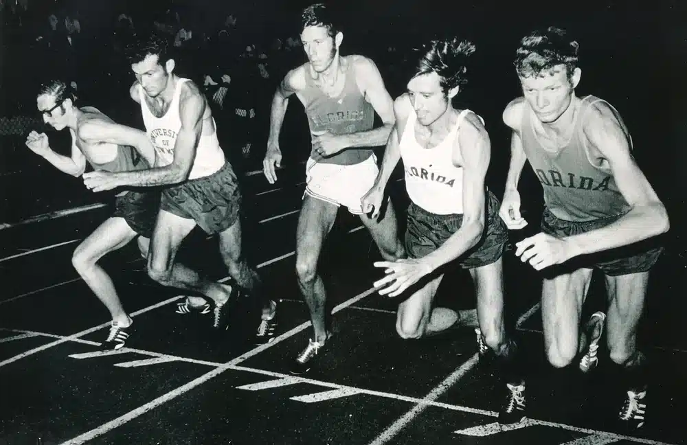 Five Florida Track Club runners in track uniforms sprinting along a track during a race.