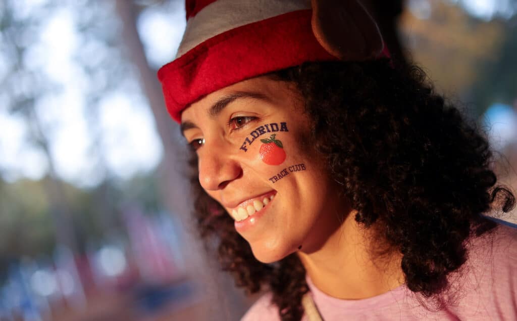 A smiling person wearing a red and white hat with a “Florida Track Club” orange sticker on their cheek, representing the Gainesville run club.